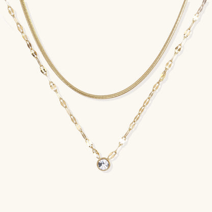 Layered gold necklace with herringbone and link chains, small bezel-set crystal pendant on cream background